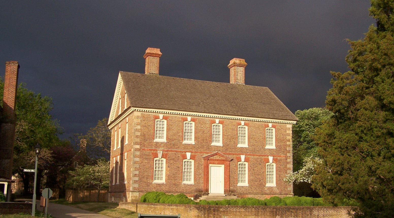 The Nelson House, a two-story brick Georgian home on Main Street in Yorktown, Virginia, lit by sun under a dark storm sky